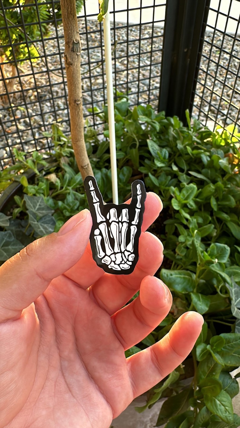 Pode incluir: Um alfinete de esmalte preto e branco em forma de m&atilde;o de esqueleto fazendo o gesto de rock and roll. O alfinete &eacute; segurado em uma m&atilde;o, com um fundo de folhagem verde e uma cerca de metal preta.