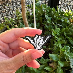 Pode incluir: Um adesivo preto e branco em forma de m&atilde;o fazendo o sinal shaka, com um design de m&atilde;o de esqueleto. O adesivo &eacute; segurado em frente a um fundo de plantas verdes e uma cerca de metal preta.