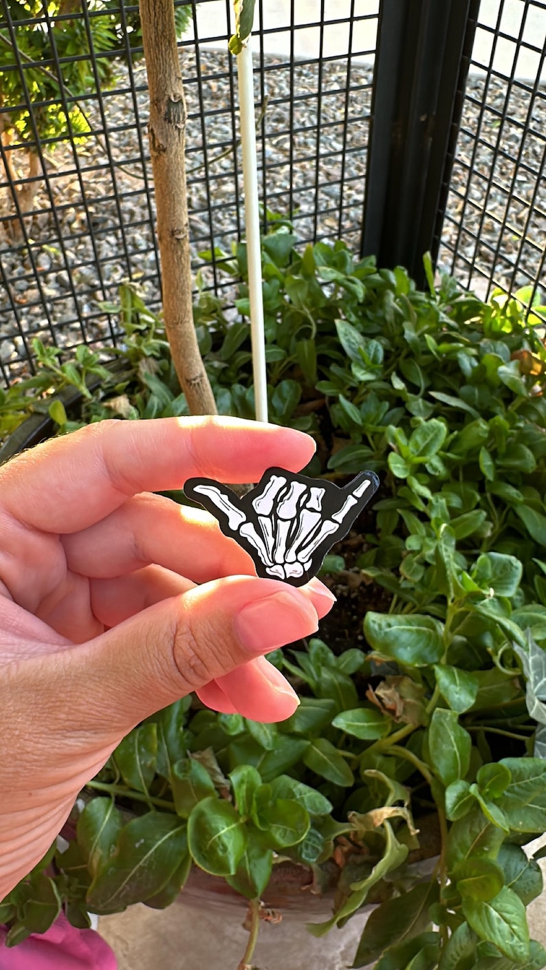 Pode incluir: Um adesivo preto e branco em forma de m&atilde;o fazendo o sinal shaka, com um design de m&atilde;o de esqueleto. O adesivo &eacute; segurado em frente a um fundo de plantas verdes e uma cerca de metal preta.