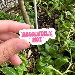 Pode incluir: Um adesivo branco com letras cor-de-rosa que diz "ABSOLUTAMENTE NÃO" com detalhes de glitter. O adesivo está sendo segurado por uma mão na frente de uma planta em vaso.