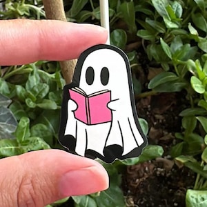 Può includere: Un adesivo di un fantasma bianco con un libro rosa nelle sue mani. Il fantasma sta tenendo il libro con entrambe le mani e sta guardando il libro. L'adesivo è su uno sfondo di pianta verde.
