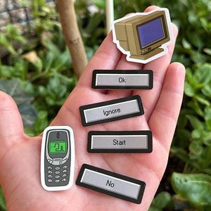 Op de afbeelding: Een hand die vijf stickers vasthoudt. De stickers zijn van een vintage computermonitor, een mobiele telefoon en drie knoppen met de tekst "Ok", "Negeren", "Starten" en "Nee".