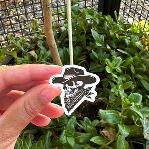 Pode incluir: Um autocolante a preto e branco com uma caveira a usar um chap&eacute;u de cowboy e uma bandana. O autocolante &eacute; segurado em frente a um fundo de plantas verdes e uma cerca de metal preta.