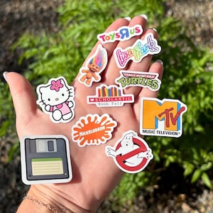 以下が含まれることがあります： ポップカルチャーのアイコンが描かれた、カラフルなステッカーのコレクション。ハローキティのステッカー、トロール人形、ニコロデオン、MTV、トイザらスのロゴが含まれています。その他、リサ・フランク、ミュータント・タートルズ、Scholastic Book Fair、フロッピーディスク、ゴーストバスターズのロゴなど。
