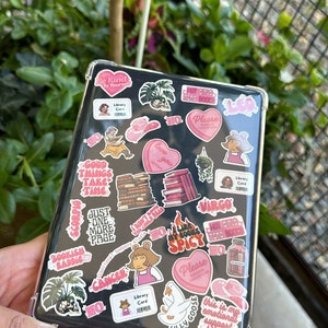 以下が含まれることがあります： ステッカーで覆われた透明な保護ケースが付いた黒いタブレット。ステッカーには、ピンクのハート、本の山、「Hot Girls Read Books」というテキストなど、さまざまなデザインが施されています。他のステッカーには「Library Card」と「Silly Goose」と書かれています。