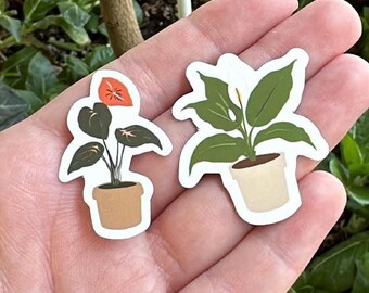 May include: Two plant stickers are held in a hand. One sticker features a plant with dark green leaves and an orange flower in a brown pot. The other sticker shows a plant with green leaves in a white pot. The background is green.