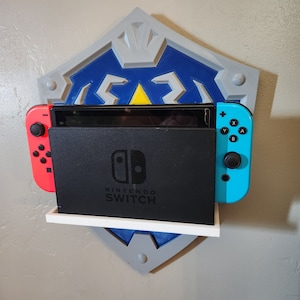 May include: A 3D printed wall mount shaped like a shield with a blue and gray design. A Nintendo Switch console is docked in the mount. The console is black with a white Nintendo Switch logo. The Joy-Con controllers are blue and red.