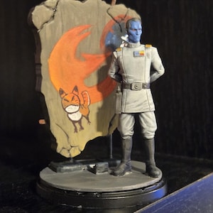 Könnte beinhalten: Figur eines blauhäutigen Humanoiden in grauer Uniform, vor einem Banner mit einem orangefarbenen Symbol und einem Cartoon-Fuchs stehend. Die Figur steht auf einem runden, grauen Sockel.