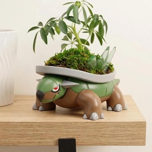 May include: A green and brown turtle-shaped planter featuring a smooth, molded plastic body with a flat, grey rim. The planter contains a small green plant nestled in dark soil and moss, with translucent, light green, pointed crystal-like accents.