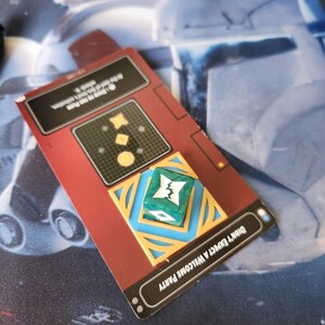 May include: A red card with the text "Don't Expect a Welcome Party" and a green and blue dice with a white symbol on it. The dice is on a gold and blue decorative square.