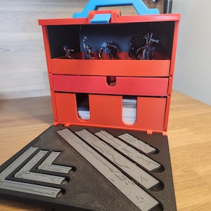 以下が含まれることがあります： 青いハンドルが付いた赤いプラスチック製の収納ケースで、ミニチュアフィギュアとゲームピースが入っています。ケースには引き出しが2つと、ゲームピースを収納するための仕切り付きの取り外し可能なトレイがあります。トレイが開いており、灰色のプラスチック製のゲームピースのセットが見えています。