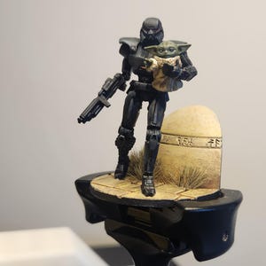 Könnte beinhalten: Eine schwarze und silberne Actionfigur hält eine kleine grüne und braune Actionfigur. Die Figuren stehen auf einem kleinen, braunen, rechteckigen Sockel.