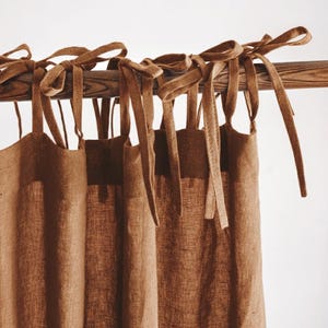 May include: Brown linen curtains with tie fastenings, suspended from a wooden pole. The curtains are a plain colour and have a textured look. The tie fastenings are made of the same material as the curtains.