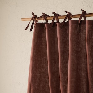 Può includere: Primo piano di tende in lino color bordeaux appese a un'asta di legno. Le tende sono legate con nastri di tessuto coordinati. Lo sfondo è un muro bianco sporco.