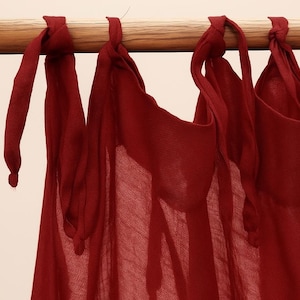 May include: Close-up of deep burgundy curtains suspended from a wooden pole. The curtains are made of a sheer material and are tied with fabric loops. The curtains are a rich red colour.