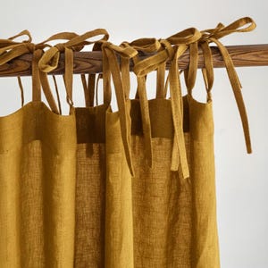 May include: Mustard-coloured linen curtains with tie closures. The curtains hang from a wooden rod. The fabric has a textured appearance, and the ties are knotted at the top. The overall aesthetic is simple and natural.