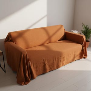 May include: A terracotta-coloured sofa cover draped over a couch. The fabric has a textured appearance, and the cover drapes over the arms and back of the sofa. The room has white walls and a wooden floor.