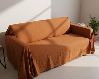 May include: A burnt orange sofa cover draped over a couch. The fabric has a textured appearance, and the cover drapes over the arms and back of the sofa. The room has white walls and a wooden floor.