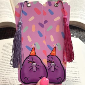 May include: Two purple bookmarks with a cartoon character holding a drink, wearing a party hat, and a colorful sprinkle pattern. Each bookmark has a tassel, one purple and one pink. The bookmarks are shown over an open book.
