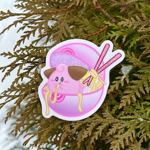 May include: A pink sticker featuring a cartoon character with brown ears and a smiling face, inside a bowl of ramen. The ramen has yellow noodles and pink chopsticks. The sticker is on a background of green branches.