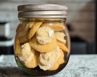 May include: A clear glass jar filled with cookies. The cookies are round, with a light yellow color and a creamy white topping. The jar has a silver lid and sits on a speckled countertop.