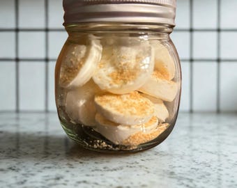 May include: A clear glass jar with a metallic lid filled with round, off-white, disc-shaped items topped with a light brown granular texture. The jar is transparent, revealing the stacked contents inside, which appear to be small, circular pieces.