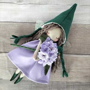 May include: A handmade fairy doll with a green pointed hat and a lavender dress. The doll has long brown hair and holds a bouquet of lavender flowers. The fairy has glittery wings and green shoes. The doll is made of fabric and is set against a light wood background.