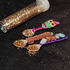 Puede incluir: Tres cucharas de plata decoradas con purpurina colorida y alambre envuelto con cuentas y piedras de colores. Las cucharas están dispuestas sobre un fondo de tela negra.