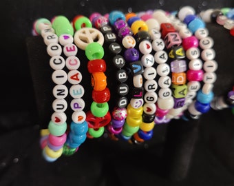 May include: A stack of colourful beaded bracelets with various words and symbols, including "Unicorn", "Peace", "Dubstep", "Grizzly", "Galaxy", "Love", and "Titi Stitch".
