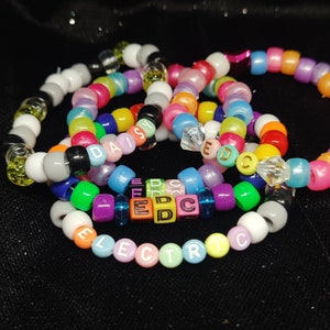 May include: Multicolored beaded bracelets with the words "Daisy" and "Electric" spelled out in colorful letter beads. The bracelets are made with small, round beads in various shades of pink, blue, green, yellow, and white.