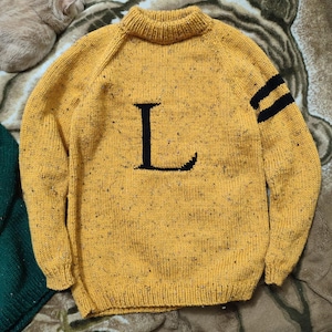 May include: Mustard yellow knit sweater with a black letter "L" on the front and two black stripes on the left sleeve. The sweater has a crew neck and a speckled texture.