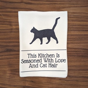 May include: White kitchen towel with a black silhouette of a cat and the text "This Kitchen Is Seasoned With Love And Cat Hair."