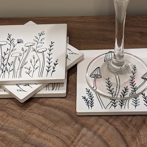 May include: Set of four white ceramic coasters with a black line drawing of mushrooms and foliage. The coasters are stacked on top of each other.