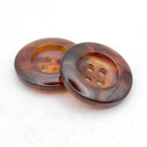 May include: Two large, round buttons with a tortoiseshell pattern. Each button has four holes for sewing. The buttons are a mix of brown and amber colours, with a glossy finish. They are set against a white background.