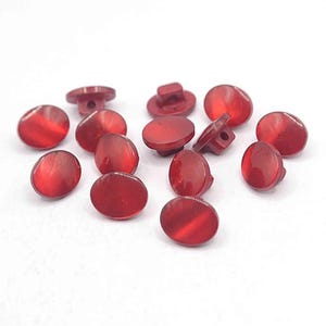 May include: A selection of red, oval buttons with a shiny finish. The buttons have a flat back with a loop for sewing. The buttons are a solid, translucent red colour and are scattered on a white surface.