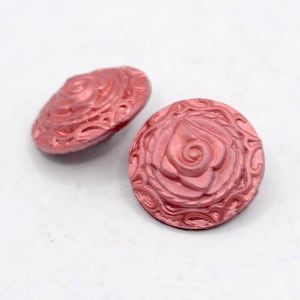 May include: Two rose-coloured, round buttons with a raised floral design. The buttons have a detailed rose and swirl pattern. The buttons are made of a shiny material and are set against a white background. These buttons are ideal for sewing and craft projects.