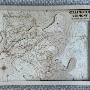 May include: Wooden topographic map of Killington, Vermont, with contour lines and a compass rose. The map includes the text "Killington Vermont" and coordinates 43.6045° N, 72.8201° W. The map is framed in a light-colored wood.