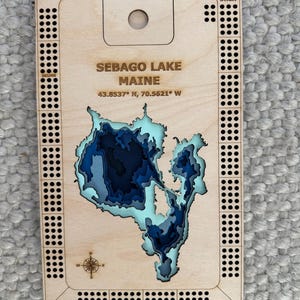 May include: A rectangular wooden cribbage board featuring a laser-cut, layered bathymetric map of Sebago Lake, Maine. The board includes the text SEBAGO LAKE MAINE 43.8537 N, 70.5621 W, a compass rose, and drilled holes for gameplay.