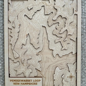 Puede incluir: Un mapa topográfico de madera del Pemigewasset Loop en New Hampshire. El mapa está cortado con láser con líneas de contorno y presenta una rosa de los vientos. El texto "PEMIGEWASSET LOOP NEW HAMPSHIRE" se muestra en la parte inferior, junto con las coordenadas.