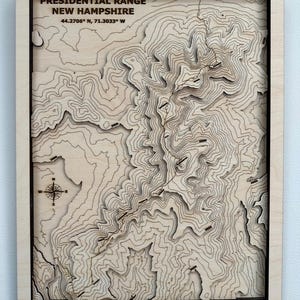 May include: Wooden topographic map of the Presidential Range in New Hampshire, with contour lines and a compass rose. The text "PRESIDENTIAL RANGE NEW HAMPSHIRE" and coordinates are visible. The map is framed in a black border.