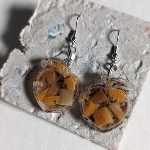 Op de afbeelding: Een paar gouden en heldere hars oorbellen in de vorm van appels. De oorbellen hebben een zilveren haak sluiting en zijn gevuld met gouden glitter en kleine stukjes goud.