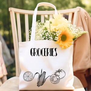 May include: A white canvas tote bag with the word "GROCERIES" in black script. The bag features line drawings of a lemon slice, corn, and a tomato. The bag is filled with yellow sunflowers and white baby's breath, and is sitting on a wooden chair.