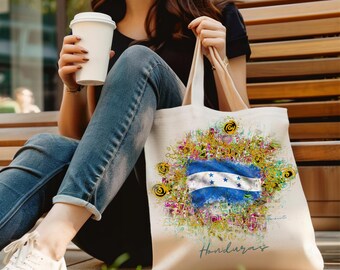 May include: A white canvas tote bag with a colorful design featuring the Honduran flag and the word "Honduras". The bag is being held by a person sitting on a wooden bench.