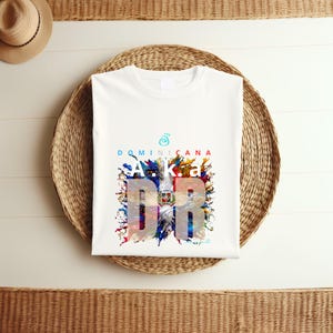 May include: White t-shirt with a colorful graphic design featuring the text "Dominicana" and "A.K.A. DR". The design incorporates a vibrant splash of colors, including blue, red, and yellow, creating a visually appealing aesthetic. The shirt is laid on a woven basket.