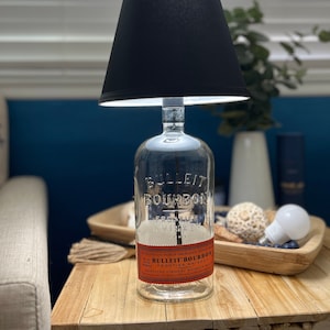 May include: A black lampshade with a clear glass bottle base. The bottle is labeled 