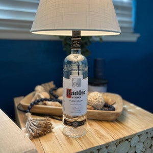 May include: A white lampshade with a woven texture sits atop a clear glass bottle of Ketel One Vodka. The bottle label reads 