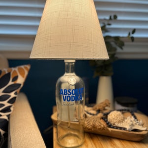 May include: A lamp made from a clear glass bottle with a white fabric lampshade. The bottle has a blue label that says 