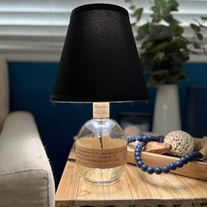 May include: A black lampshade with a clear glass bottle base. The bottle is labeled "Blanton's" and has a brown paper label with text. The lamp is sitting on a wooden table with a blue beaded garland and other decorative items.