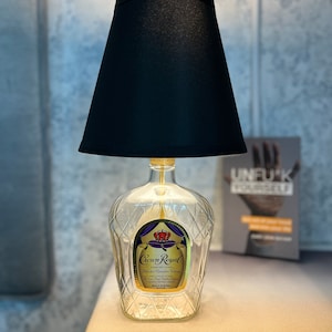 May include: A lamp made from a repurposed Crown Royal whiskey bottle. The clear glass bottle base features a gold label with purple and gold accents. A black lampshade sits atop the bottle, providing a contrasting element to the clear glass.