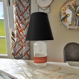 May include: A black lampshade with a clear glass bottle base. The bottle has a label that reads 